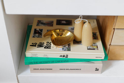 A pile of books on a shelf. On top of the books is a gold ashtray and two cream candles. 