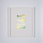 A framed print of a white page with metallic gold letters on it. This is a limited edition artwork by Rory Pilgrim. The text says Pick Up The Phone and Sound the alarm.