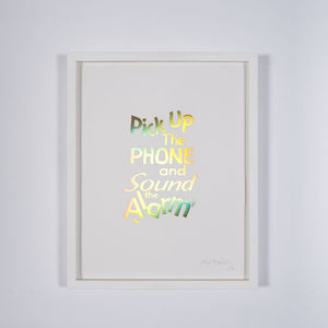 A framed print of a white page with metallic gold letters on it. This is a limited edition artwork by Rory Pilgrim. The text says Pick Up The Phone and Sound the alarm.