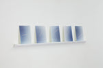 5 postcards on a white shelf. The postcards are images of shooting stars across a blue sky. The work is a print by artist Ann Veronica Janssens.