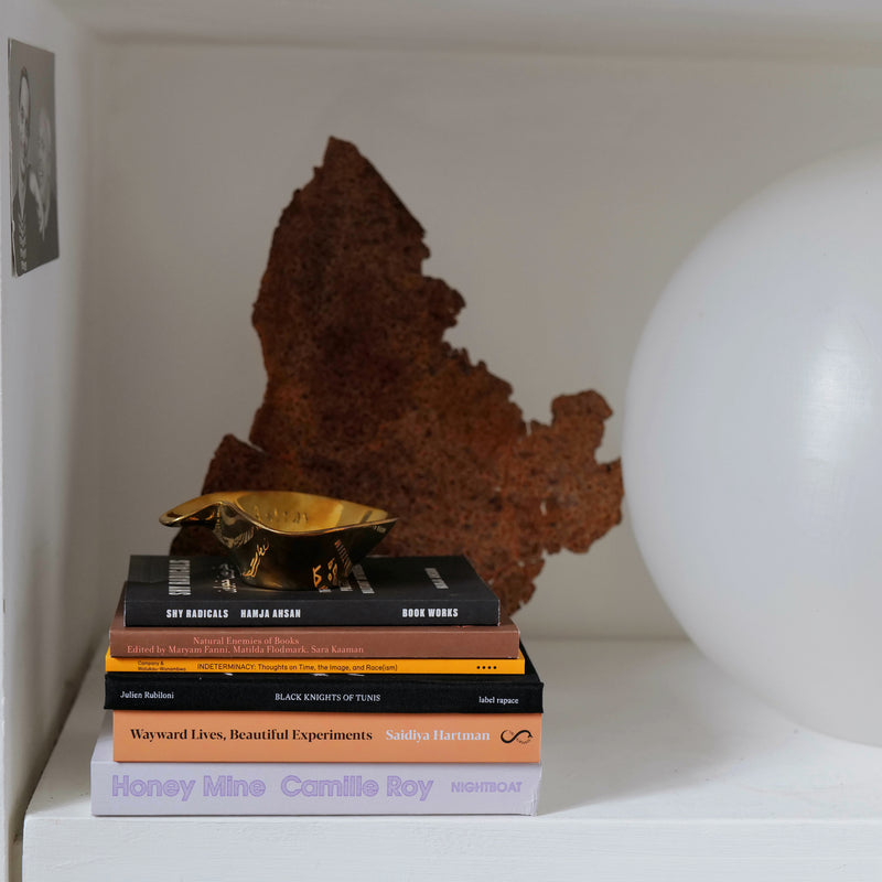 A pile of books on a white shelf. On top of the books is a gold sculptural ashtray.