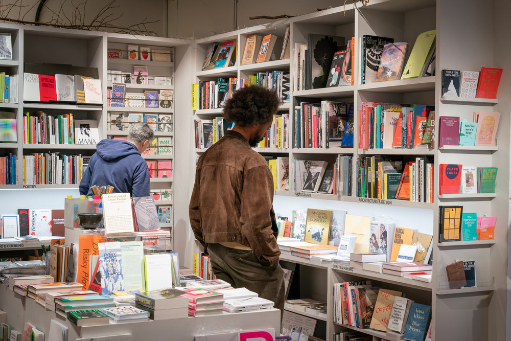 Two people stand in a bookshop and look at colourful books on shelves. 