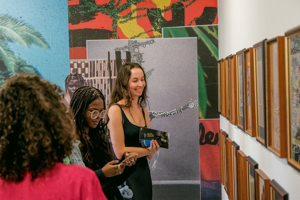 Two people stand in an art gallery looking at art. There are lots of framed pictures on the wall and a large collage on one wall.