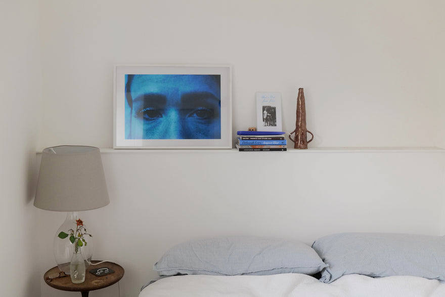 A framed artist print, vase and books sit on shelf in a bedroom.