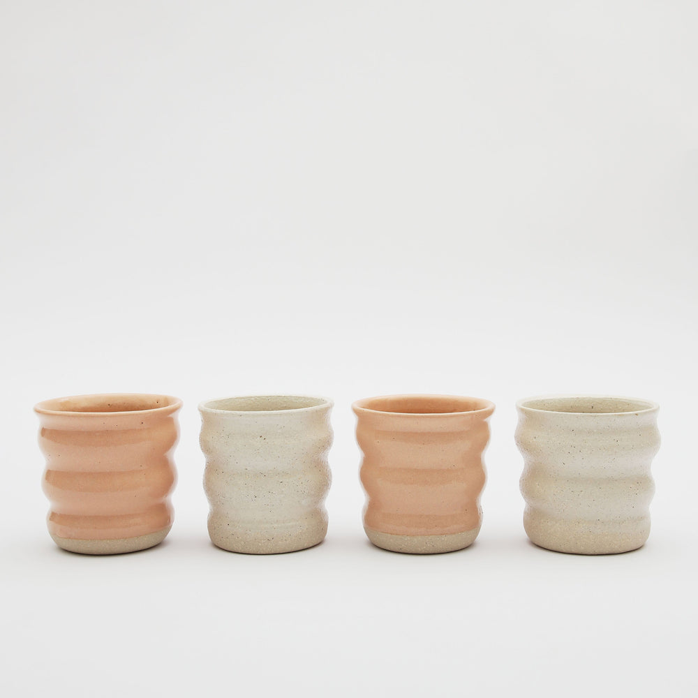 Four ceramic mugs by Gioz Ceramics. The mugs have no handles and are glazed in peach and an off white stone. 