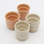Four ceramic mugs by Gioz Ceramics. The mugs have no handles and are glazed in peach and an off white stone.