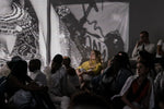 People sit on the ground in a dark room. There are black and white photography projections on the wall. One woman with blonde hair and a yellow top is being bathed in light from the projections. 