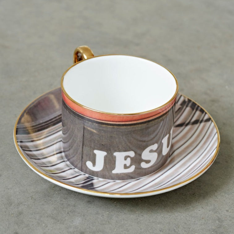 A china teacup and saucer by the artist Rene Matic. The teacup is printed with a photograph with the word Jesus on it.