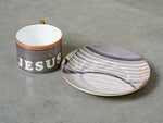 A china teacup and saucer by artist Rene Matic. The teacup has been printed with the word Jesus. 