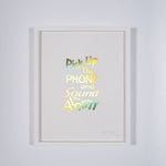 A framed print of a white page with metallic gold letters on it. This is a limited edition artwork by Rory Pilgrim. The text says Pick Up The Phone and Sound the alarm. 