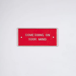 A limited artist edition by Peter Liversidge. This is an aluminium red plaque with white text that says Something on Your Mind