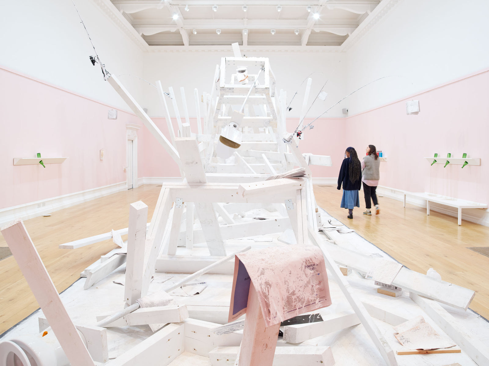 Two people walk through an art gallery and look at a large installation by artist Pope.L. The installation is made of white wood. The walls of the gallery are pink.