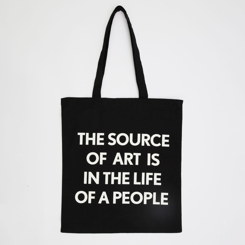 A black tote bag with white writing. The text says 'The Source of Art is in the Life of the People'.