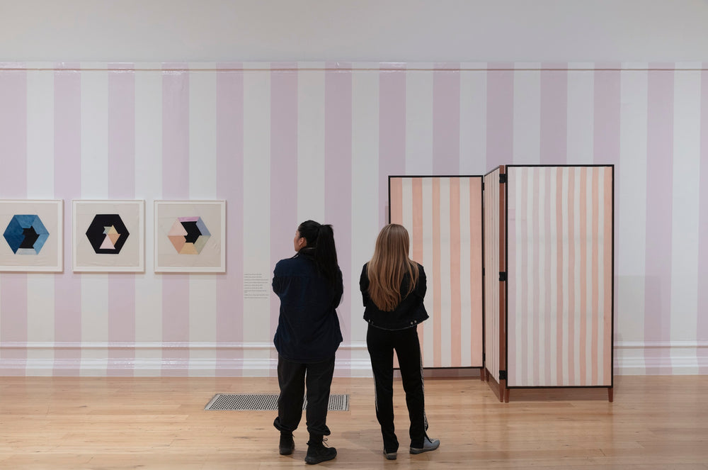 Two people look at art in an art gallery. The walls are painted with pink stripes. 