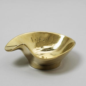 A gold ashtray by artist Becky Beasley on a grey background with the word astray engraved on the inside.