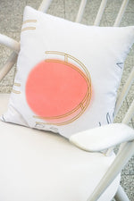 A square cushion on a white chair. The cushion is white with an orange circular pattern and yellow ladder-like illustrations.