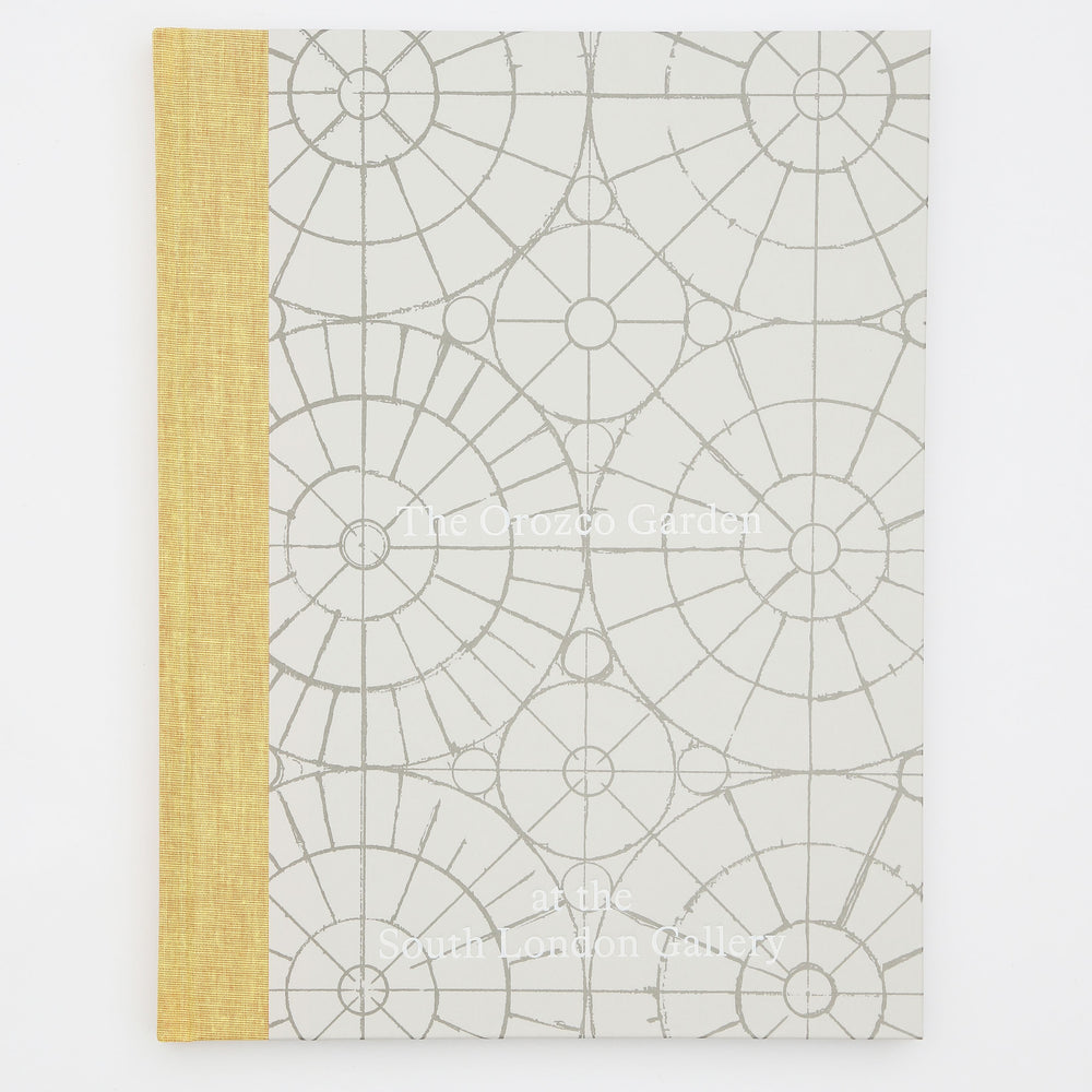 A book titled The Orozco Garden at the South London Gallery. The cover of the book is grey with a yellow spine. There are geometric patterns and circles underneath the text on the front.