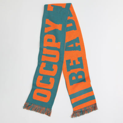 A bright orange and green football scarf with writing on it. Made by artist duo Yara and Davina. 